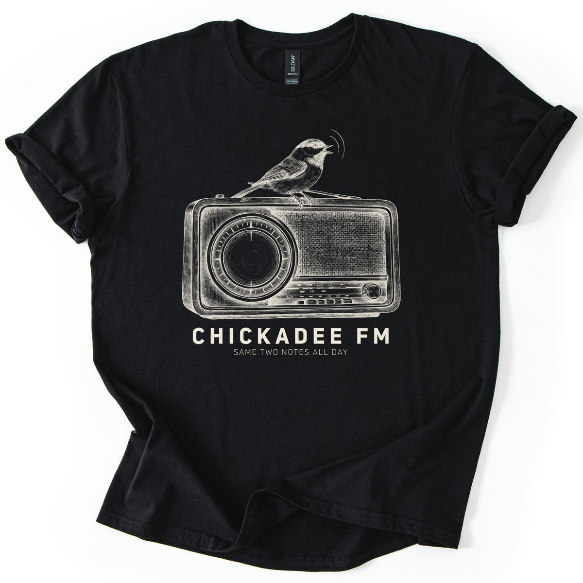 Chickadee FM Comfort Colors T-shirt in Black, laid flat. A sketched chickadee sings from atop a vintage radio above the words CHICKADEE FM and SAME TWO NOTES ALL DAY, printed in cream. The shirt has rolled sleeves and lies on a white background.