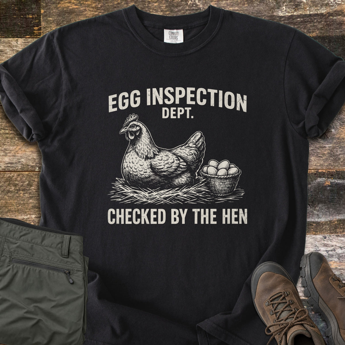 Egg Inspection Dept Comfort Colors T-shirt in Black, laid flat. A sketched hen sits on straw beside a basket of eggs, with Egg Inspection Dept. above and Checked By The Hen below in cream. Olive cargo pants and brown hiking boots lie on a rustic wood floor.