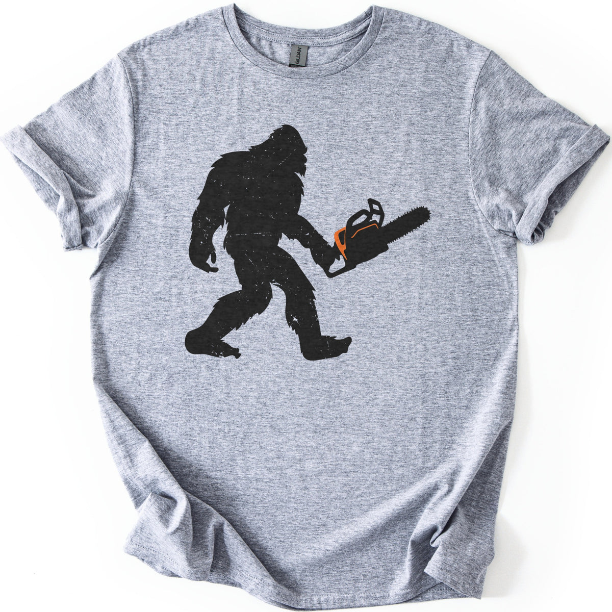 Lumber Squatch Comfort Colors T-shirt in Sport Grey, laid flat. A distressed black silhouette of Bigfoot walking mid-stride while holding a chainsaw with an orange accent, with no words printed. The shirt has cuffed sleeves and lies on a white background.