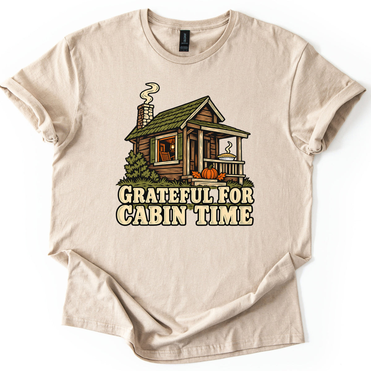 Grateful For Cabin Time Comfort Colors T-shirt in Sand, laid flat. A wooden cabin with a smoking chimney, a rocking chair seen through the lit window, a pie on the porch and a pumpkin with autumn leaves sits above the words Grateful For Cabin Time. Shown on white.