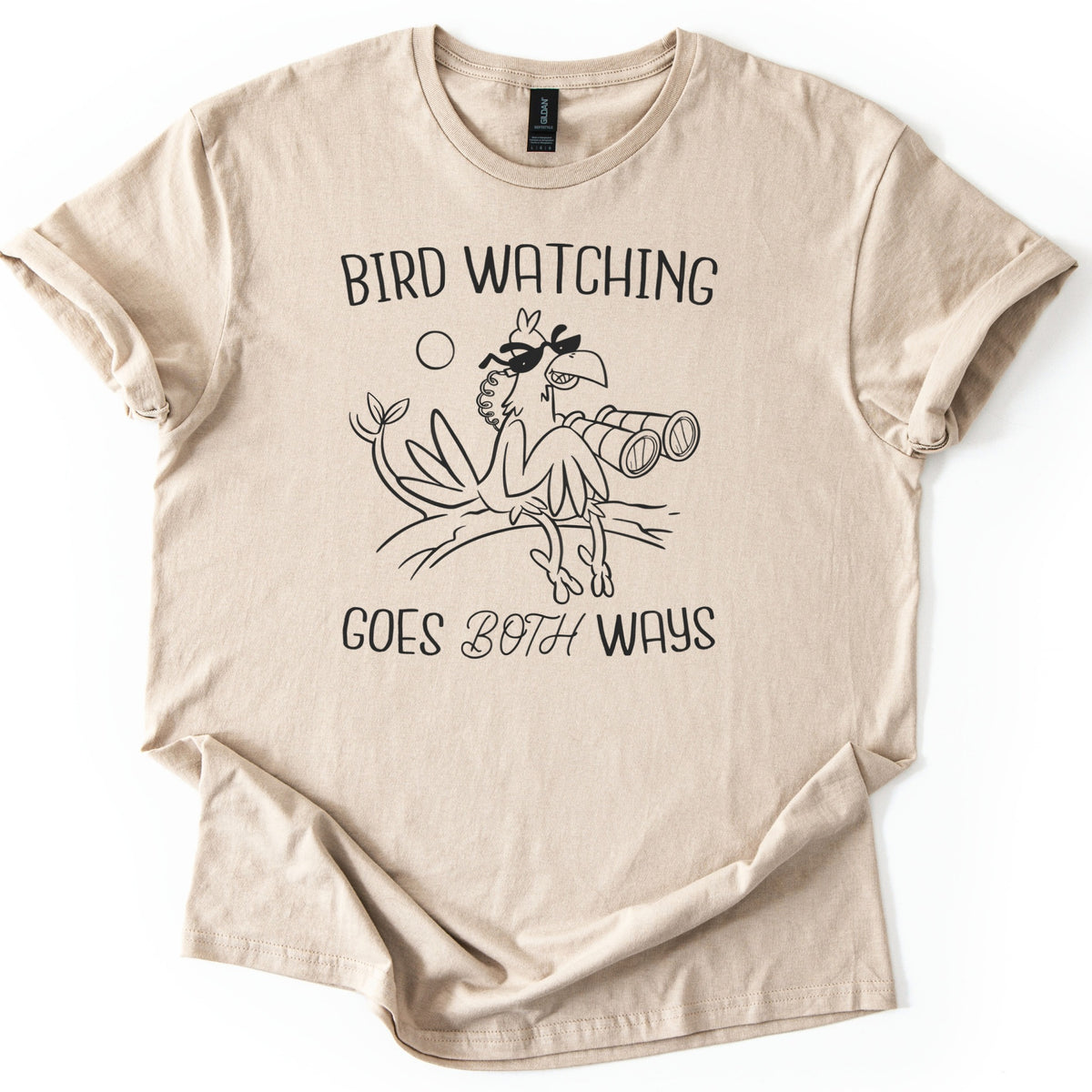 Birds Watch Back Birder Comfort Colors T-shirt in Sand, laid flat. Black line art of a cartoon bird in sunglasses perched on a branch beside a pair of binoculars, with a small sun, between the words Bird Watching Goes Both Ways. Sleeves rolled, on a white background.
