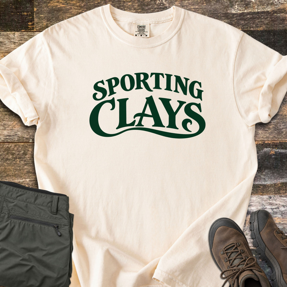 Sporting Clays Comfort Colors T-shirt in Ivory, laid flat. Dark green retro lettering reads Sporting Clays, with a swash curving under the word Clays. Sleeves are rolled on a rustic wood floor beside folded olive hiking pants and brown leather hiking boots.