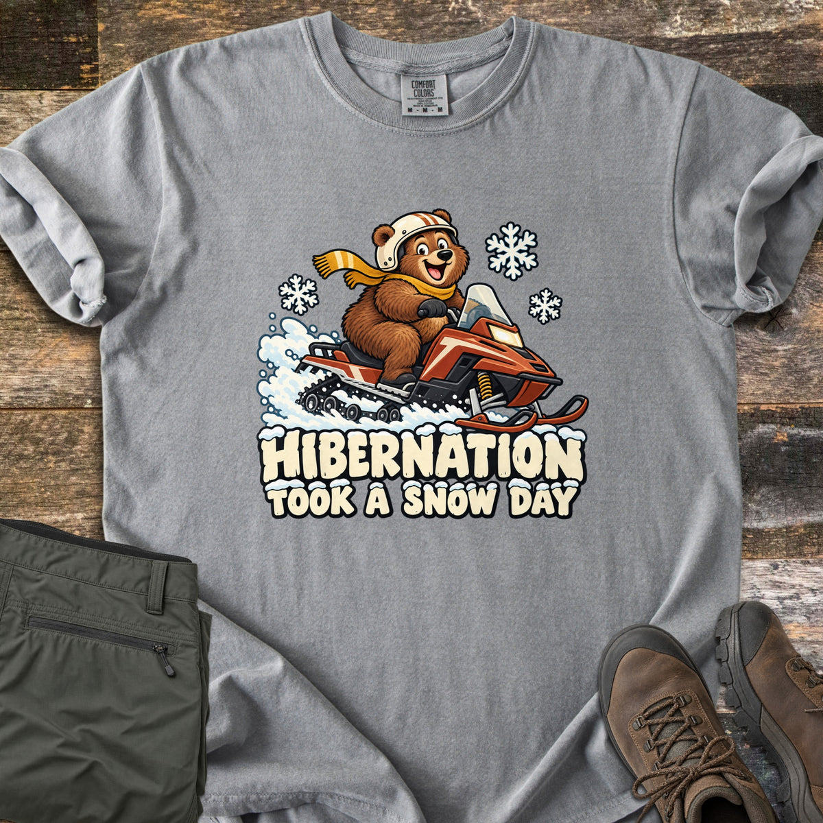 Hibernation Took a Snow Day Comfort Colors T-shirt in Grey, laid flat. A smiling cartoon bear in a helmet and yellow scarf kicks up snow on a red snowmobile above the words Hibernation Took a Snow Day. Folded olive pants and brown boots rest on dark wood planks.