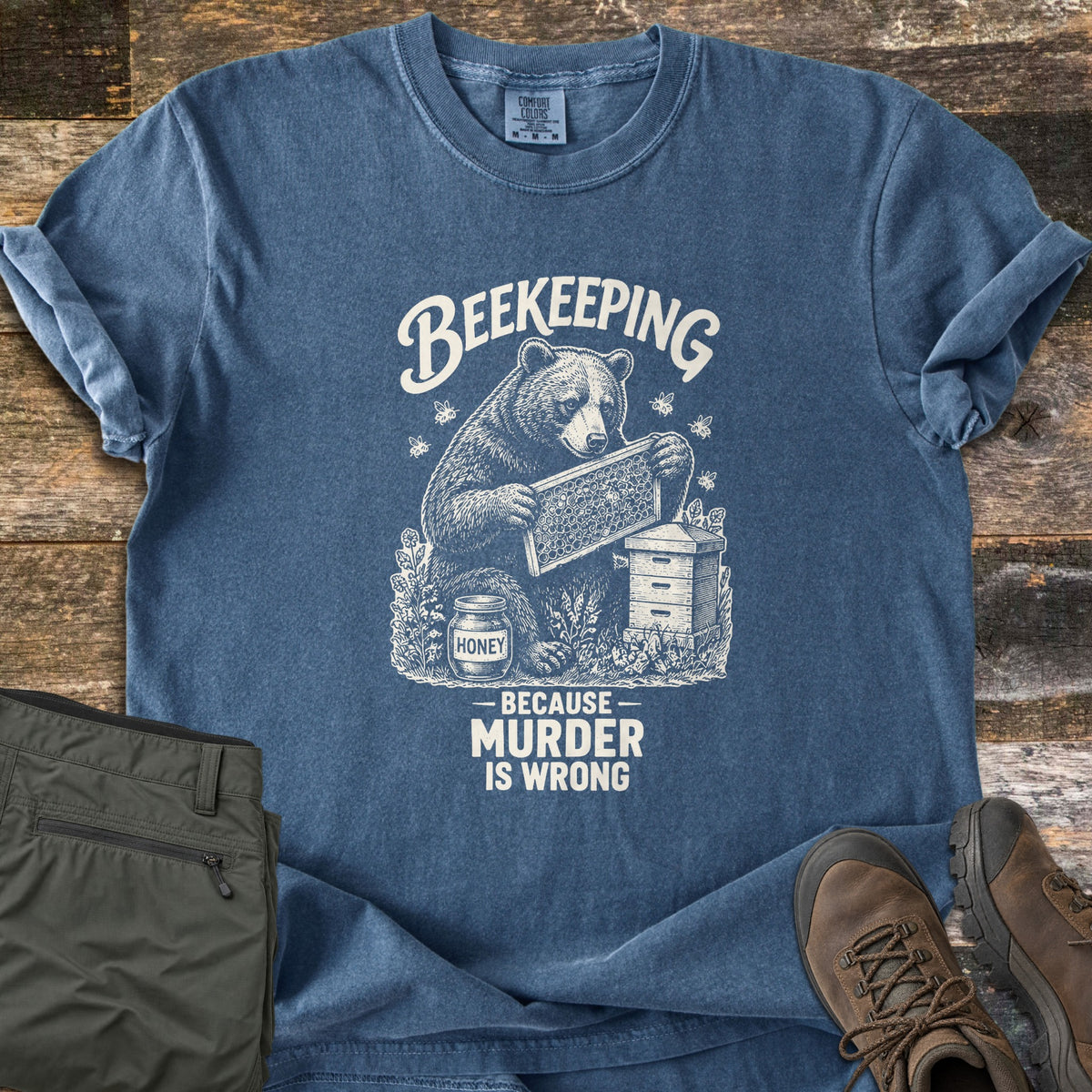 Beekeeping Because Murder Is Wrong Comfort Colors T-shirt in Denim, laid flat. Cream ink artwork shows a bear holding a honeycomb frame beside a beehive and a jar labeled Honey, with bees around it, under the word Beekeeping and above Because Murder Is Wrong. Olive pants and brown hiking boots lie on wood.