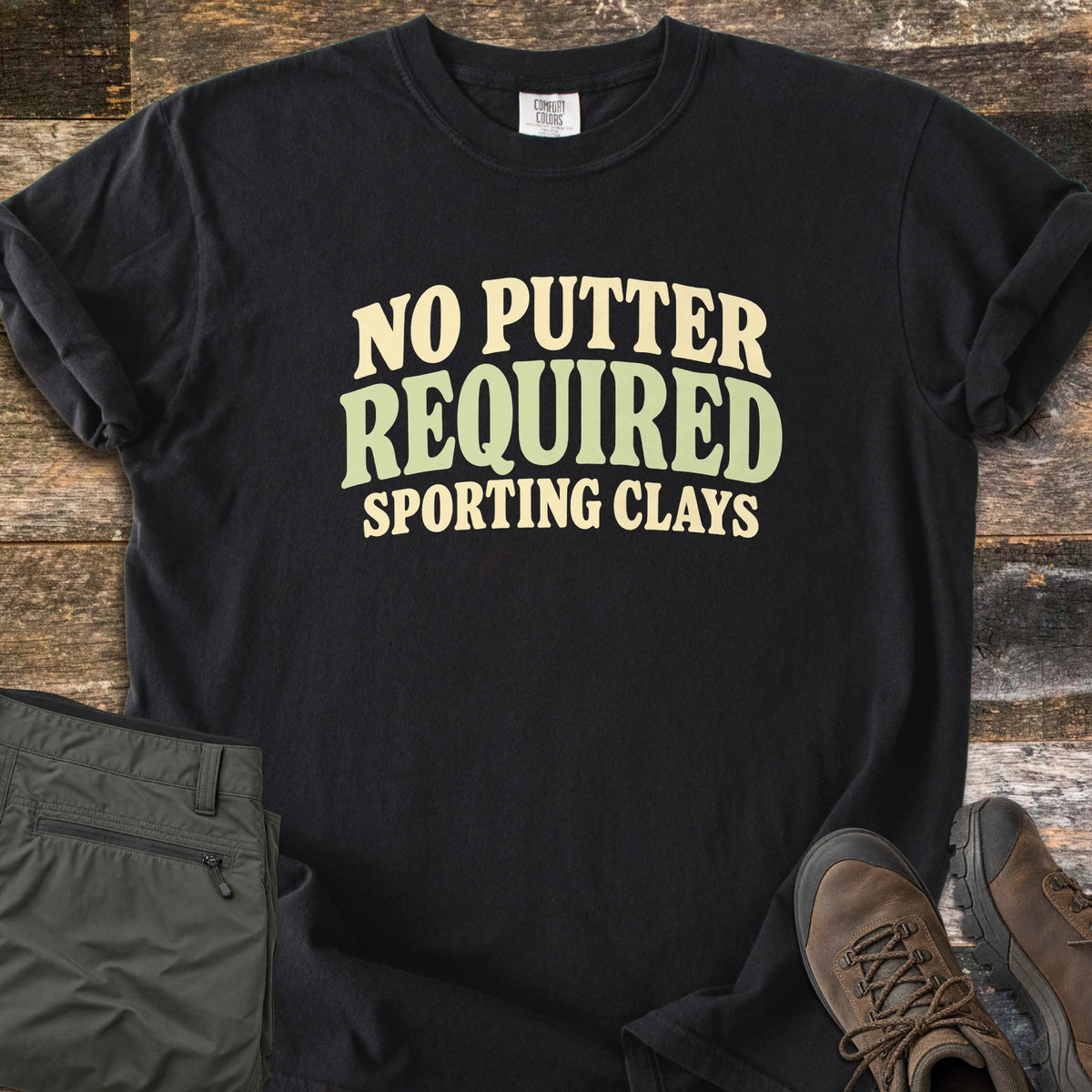 No Putter Required Comfort Colors T-shirt in Black, laid flat. Wavy block letters read No Putter Required in cream and sage green, with Sporting Clays below. Sleeves are rolled on a rustic wood surface beside folded olive cargo pants and brown leather hiking boots.