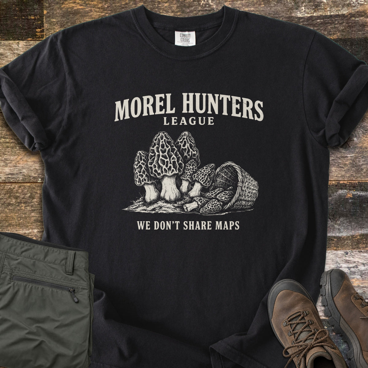 Morel Hunters League Comfort Colors T-shirt in Black, laid flat. Cream sketch of morels next to a tipped wicker basket spilling morels, under Morel Hunters League and above We Don't Share Maps. Sleeves rolled, on weathered wood with olive hiking pants and brown hiking boots.