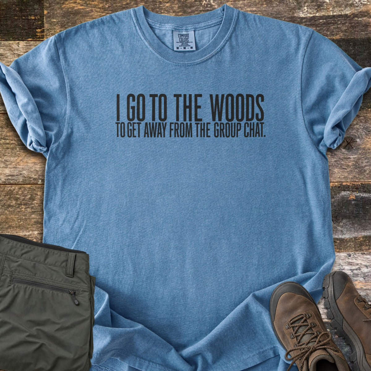 Away From The Group Chat Comfort Colors T-shirt in Blue Jean, laid flat. Black block letters read I GO TO THE WOODS above TO GET AWAY FROM THE GROUP CHAT. The shirt has rolled sleeves and lies on weathered wood boards beside folded olive green pants and brown hiking boots.