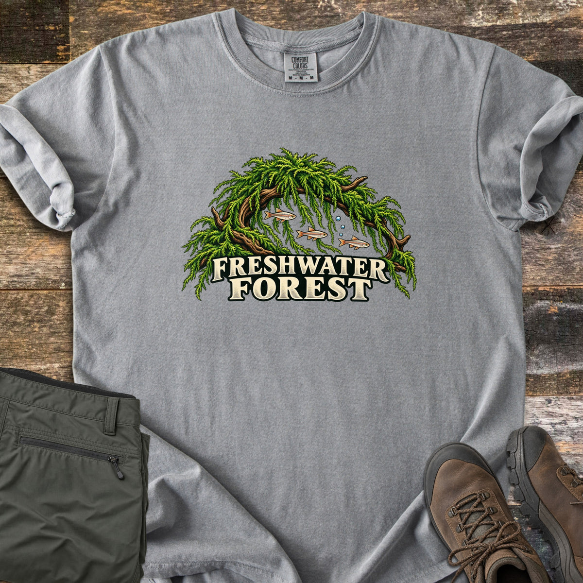 Freshwater Forest Comfort Colors T-shirt in Grey, laid flat. A mossy driftwood arch with three small fish and bubbles beneath it sits above the words Freshwater Forest. The shirt rests on weathered planks with folded olive pants and brown hiking boots.