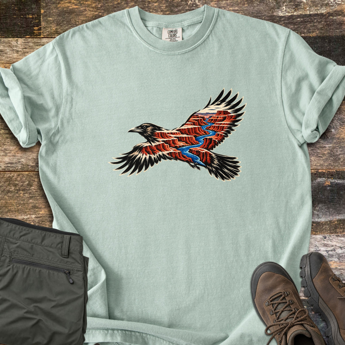Red Canyon Raven Comfort Colors T-shirt in Bay, laid flat. A black raven with spread wings, filled with layered red canyon cliffs cut by a blue river. The pale sage shirt has rolled sleeves, on rustic wood with folded olive pants and brown hiking boots.