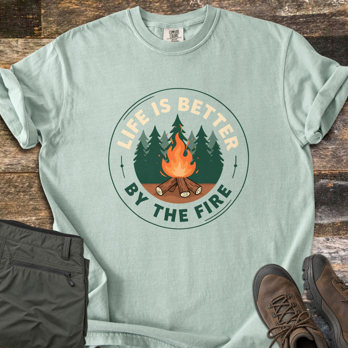 Life Is Better By The Fire Badge Comfort Colors T-shirt in Bay, laid flat. A round badge shows a campfire of crossed logs before pine trees, with LIFE IS BETTER arched above and BY THE FIRE below. Olive hiking pants and brown hiking boots sit beside it on weathered wood planks.