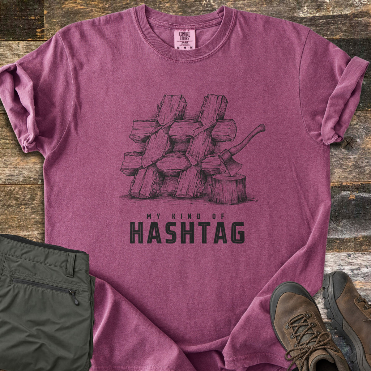 My Kind Of Hashtag Comfort Colors T-shirt in Berry, laid flat. A sketched stack of split firewood logs arranged in a hashtag shape, with an axe in a stump, above the words My Kind Of Hashtag. Olive hiking pants and brown hiking boots lie beside it on weathered wood planks.