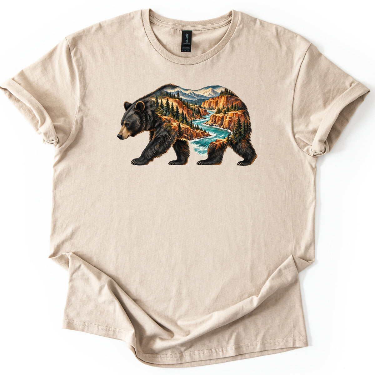 Bear River Canyon Comfort Colors T-shirt in Sand, laid flat. A walking black bear silhouette filled with a canyon scene: a turquoise river winding between orange cliffs, pine trees, and snowy mountains. The shirt lies on a white background with sleeves rolled.