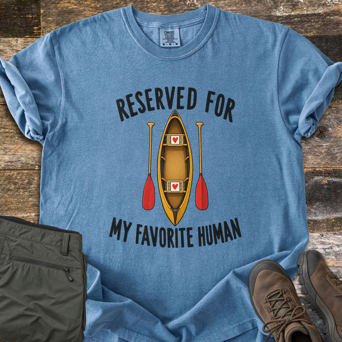 Reserved For My Favorite Human Comfort Colors T-shirt in Blue Jean, laid flat. A yellow canoe seen from above with a red heart on each seat sits between two red-bladed paddles, under the words Reserved For and above My Favorite Human. Olive pants and brown hiking boots lie beside it on wood planks.