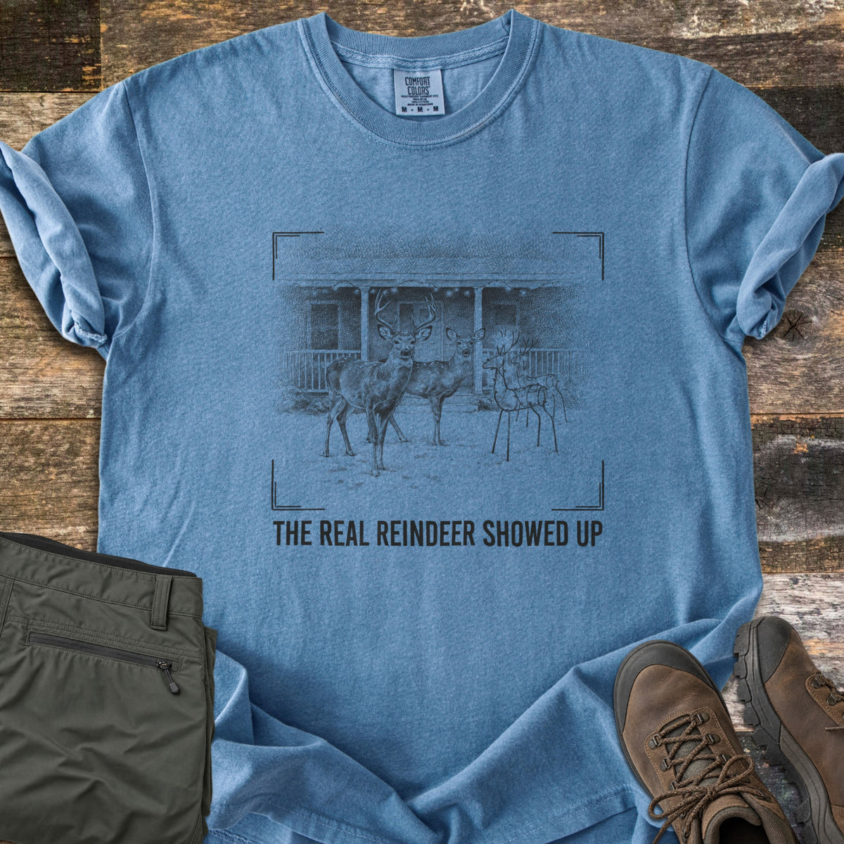 The Real Reindeer Showed Up Comfort Colors T-shirt in Blue Jean, laid flat. A sketch of a buck and doe standing in snow next to a wire reindeer lawn decoration in front of a porch, inside camera viewfinder corners, above the words The Real Reindeer Showed Up. Olive pants and brown boots on wood.