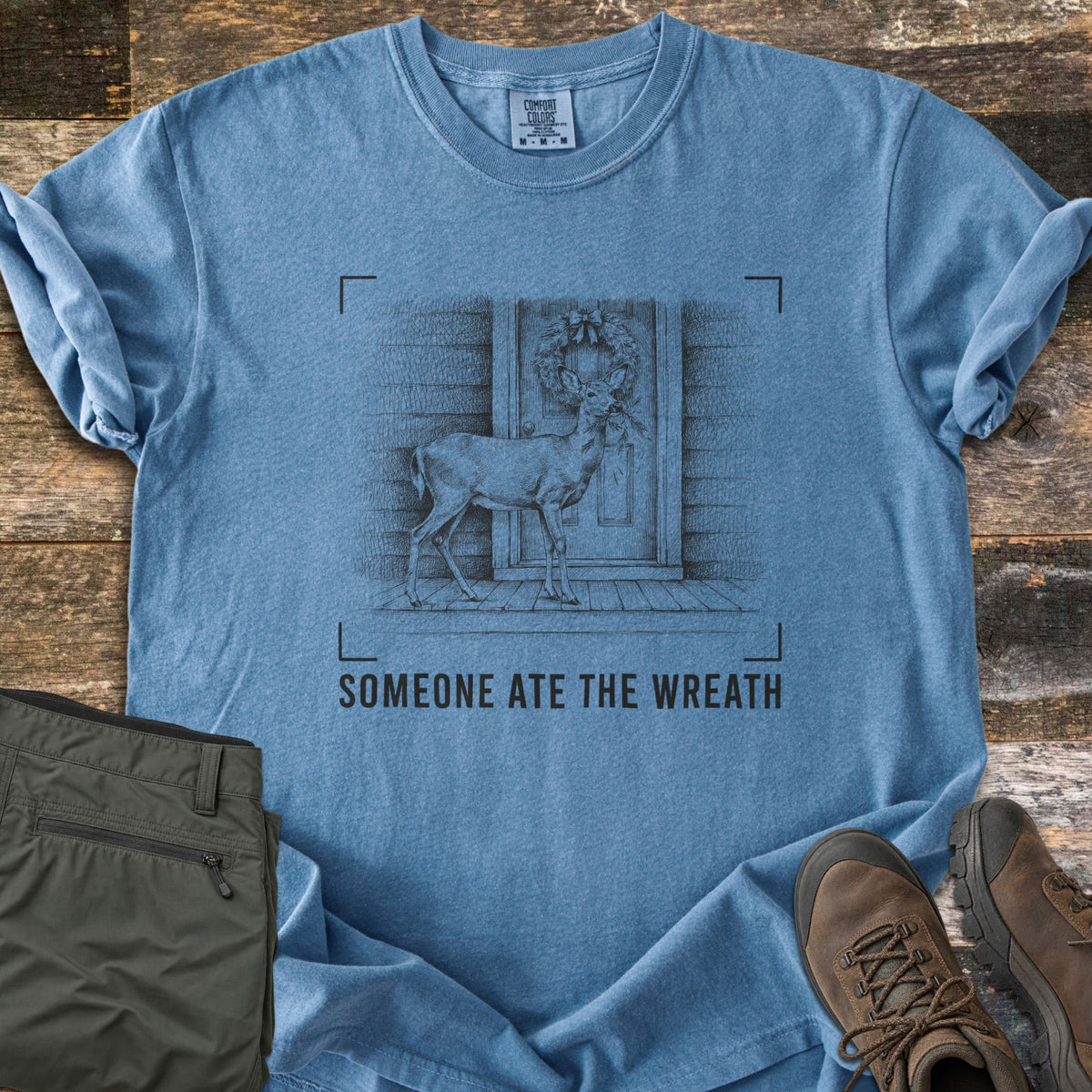 Someone Ate The Wreath Comfort Colors T-shirt in Blue Jean, laid flat. A sketched deer nibbles a wreath on a cabin's front door, framed by corner marks above the words Someone Ate The Wreath. Shown on rustic wood planks with folded olive pants and brown hiking boots.