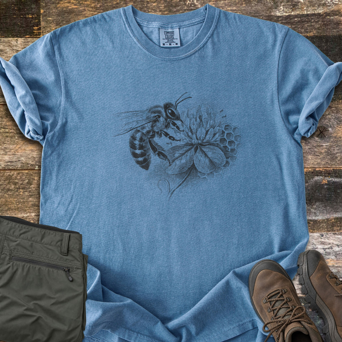 Honeybee On Clover Comfort Colors T-shirt in Blue Jean, laid flat. A detailed black engraving-style illustration of a honeybee on a clover blossom with honeycomb cells behind it, no words. Sleeves rolled, on weathered wood planks beside olive hiking pants and brown hiking boots.