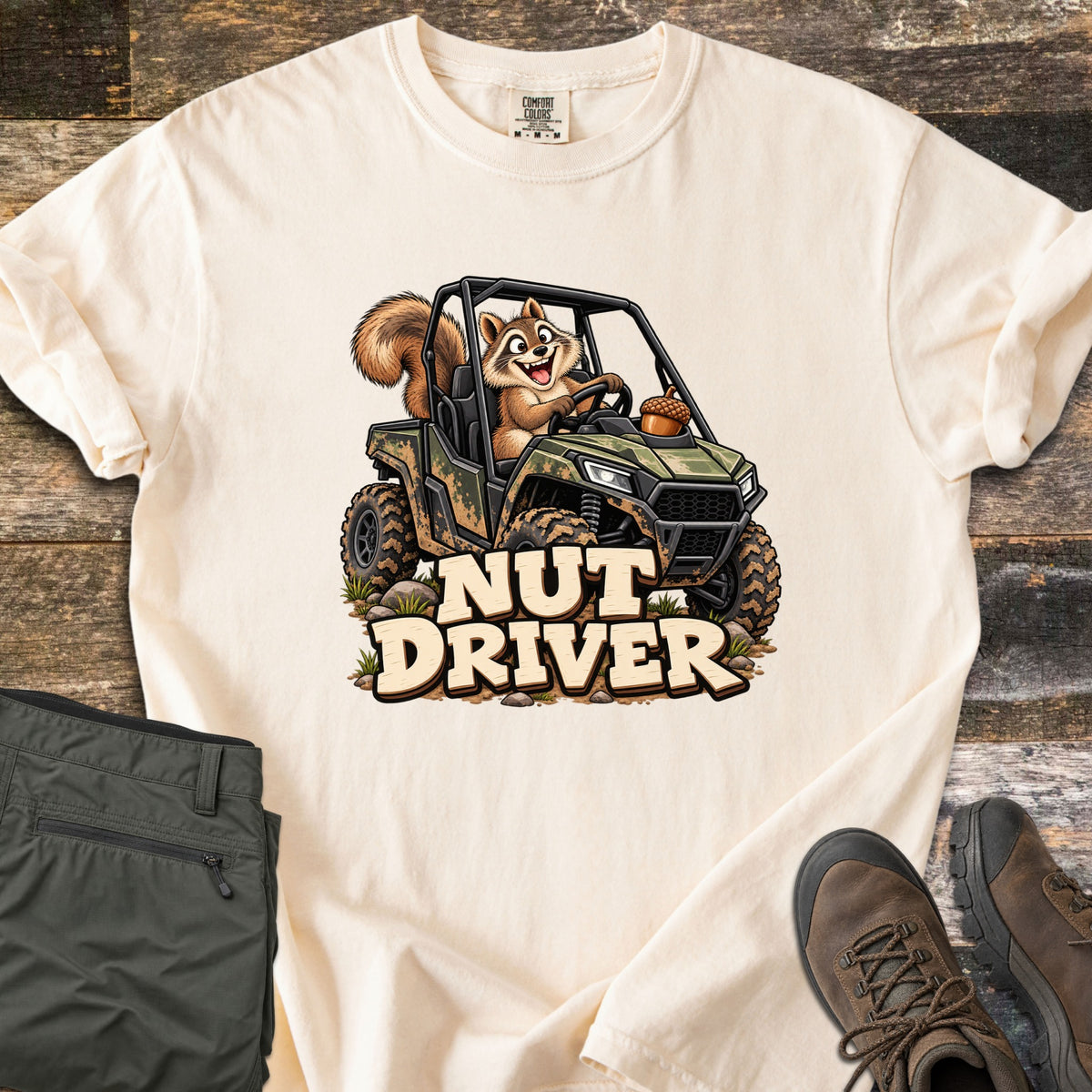 Nut Driver Comfort Colors T-shirt in Ivory, laid flat. A cartoon squirrel grins while driving a muddy camo side-by-side with an acorn on the hood, above the words Nut Driver. Olive cargo pants and brown hiking boots sit beside it on weathered wood.