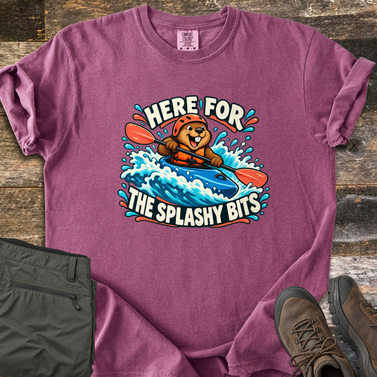 Splashy Bits Comfort Colors T-shirt in Berry, laid flat. A cartoon beaver in an orange helmet and life vest paddles a blue kayak through whitewater, framed by the words Here For The Splashy Bits. Olive pants and brown hiking boots lie beside it on weathered wood.