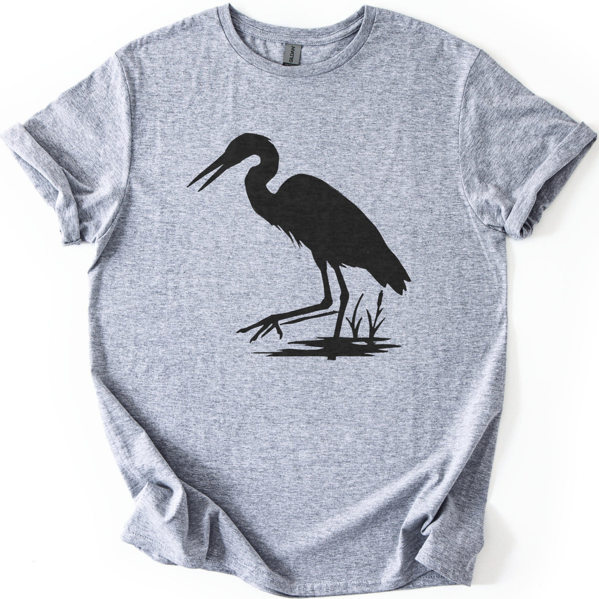 Great Blue Heron Comfort Colors T-shirt in Sport Grey, laid flat. A black silhouette of a heron wading in shallow water beside cattails, beak open and one foot reaching forward. The shirt lies on a white surface with cuffed sleeves.