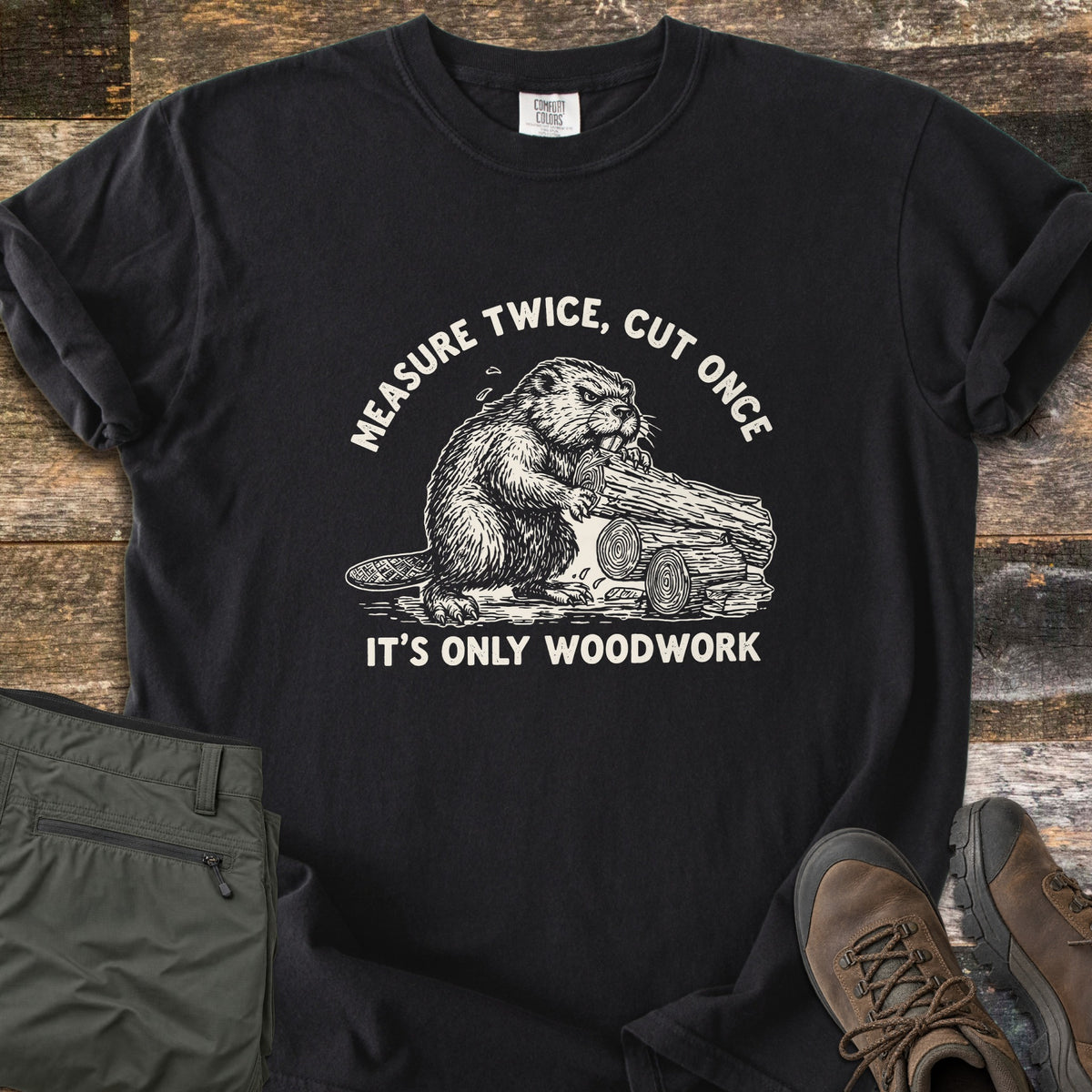 Measure Twice Beaver Comfort Colors T-shirt in Black, laid flat. A sketched beaver gnawing a log sits between the words Measure Twice, Cut Once and It's Only Woodwork. The shirt lies on weathered wood planks beside folded olive hiking pants and brown hiking boots.