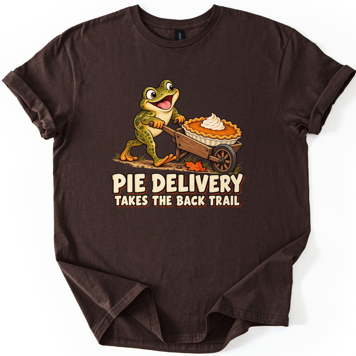Pie Delivery Takes the Back Trail Comfort Colors T-shirt in Dark Chocolate, laid flat. A happy cartoon frog wheels a pumpkin pie topped with whipped cream in a wooden wheelbarrow down a dirt trail, above the words Pie Delivery Takes the Back Trail. Rolled sleeves on a white background.