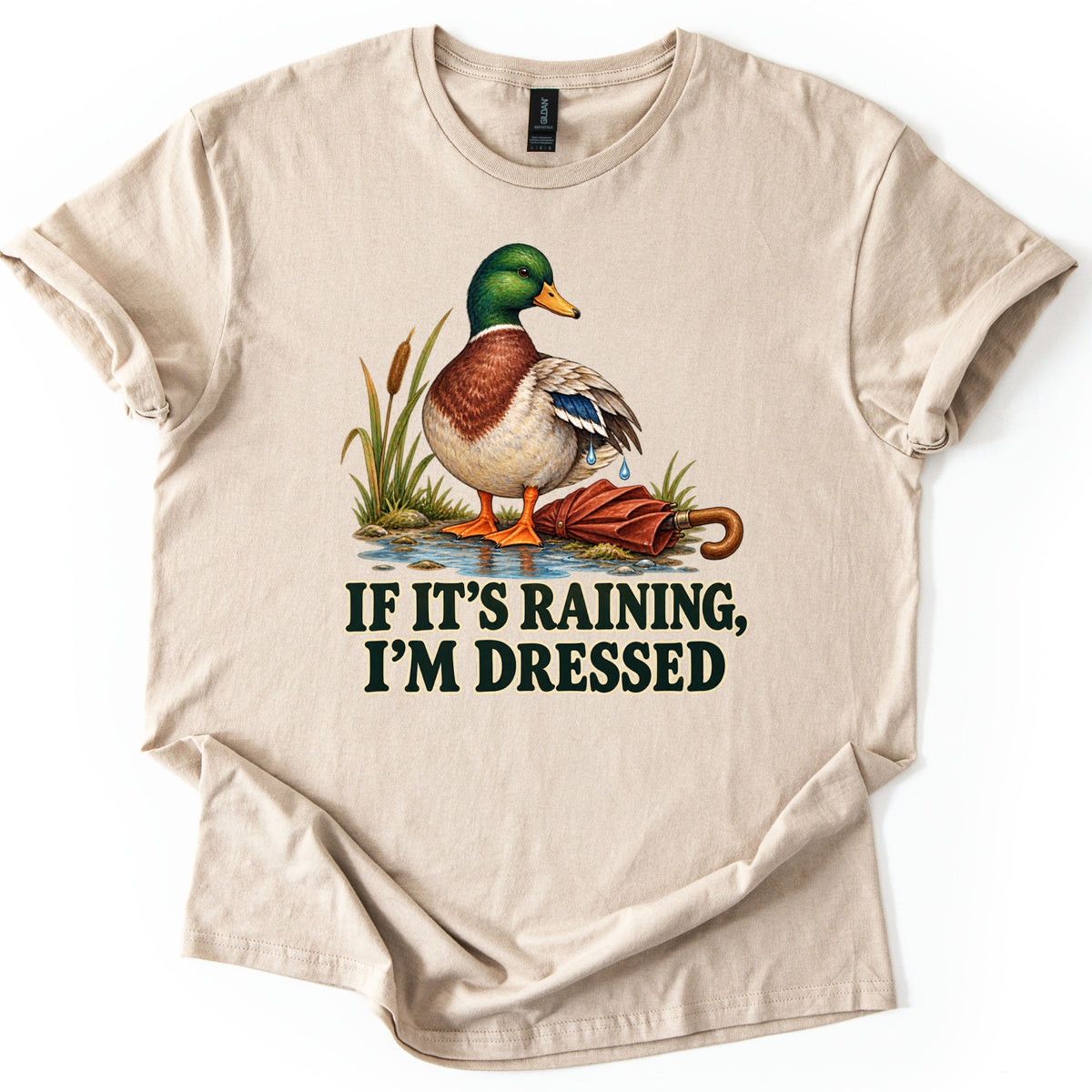 If It's Raining, I'm Dressed Comfort Colors T-shirt in Sand, laid flat. A mallard duck with water dripping off its wing stands in a puddle by cattails beside a closed red umbrella, above the words IF IT'S RAINING, I'M DRESSED in dark green. Cuffed sleeves, white background.
