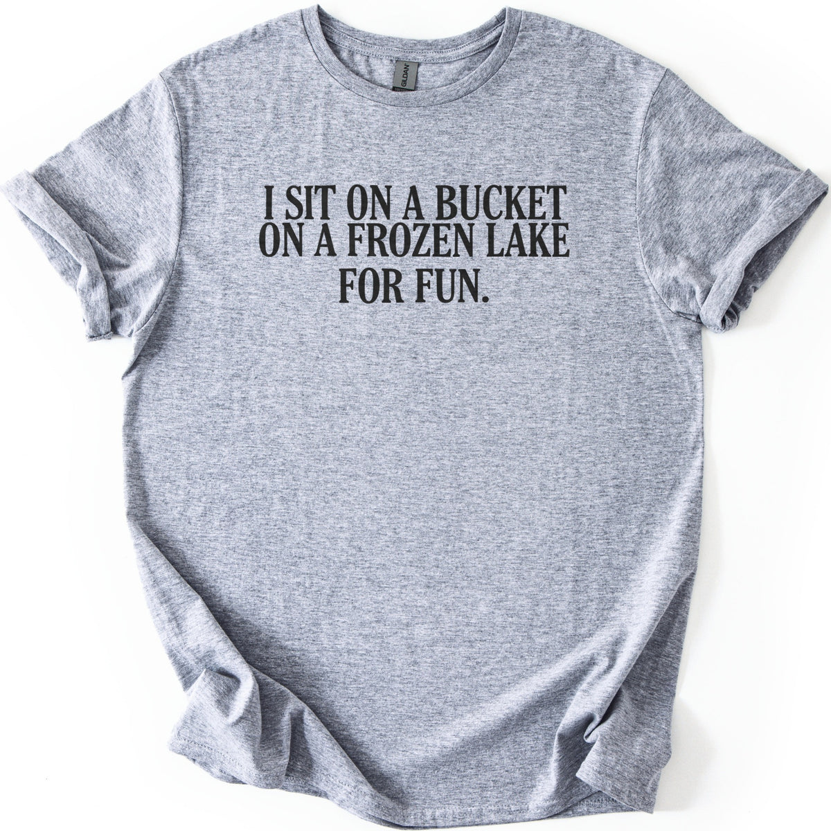 Bucket On A Frozen Lake Comfort Colors T-shirt in Sport Grey, laid flat. Black serif capital letters across the chest read I SIT ON A BUCKET ON A FROZEN LAKE FOR FUN. The shirt lies on a white surface with its sleeves rolled and the hem loosely folded.