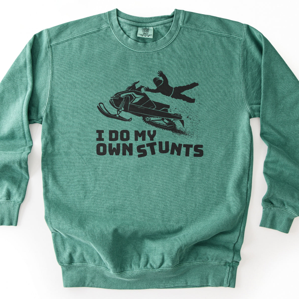 I Do My Own Stunts Comfort Colors crewneck sweatshirt in Light Green, laid flat. A black print shows a helmeted rider flying off an airborne snowmobile with snow kicking up behind, above the words I DO MY OWN STUNTS. The sweatshirt lies on a white surface.