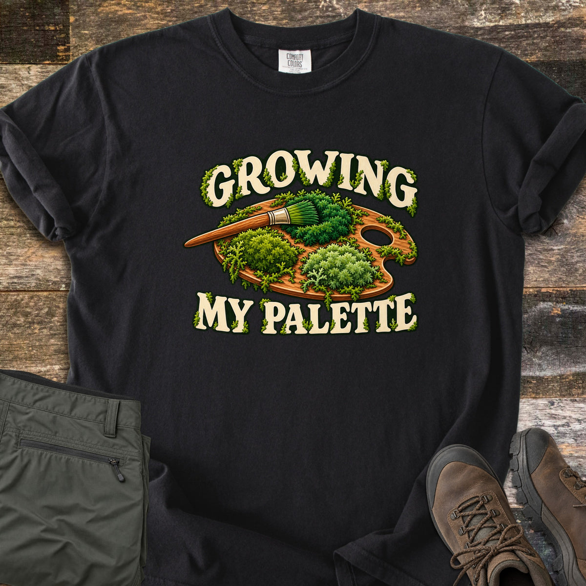 Growing My Palette Comfort Colors T-shirt in Black, laid flat. A wooden painter's palette covered in green moss, with a brush whose bristles are moss, between the words Growing My Palette in cream letters. Olive pants and brown hiking boots lie beside it on weathered wood.