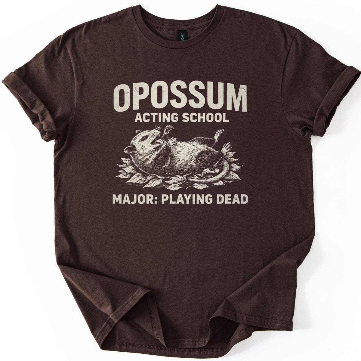 Opossum Acting School Comfort Colors T-shirt in Dark Chocolate, laid flat. A cream sketch of an opossum lying on its back on a bed of leaves, mouth open, sits under the words OPOSSUM ACTING SCHOOL and above MAJOR: PLAYING DEAD. The sleeves are rolled on a white background.