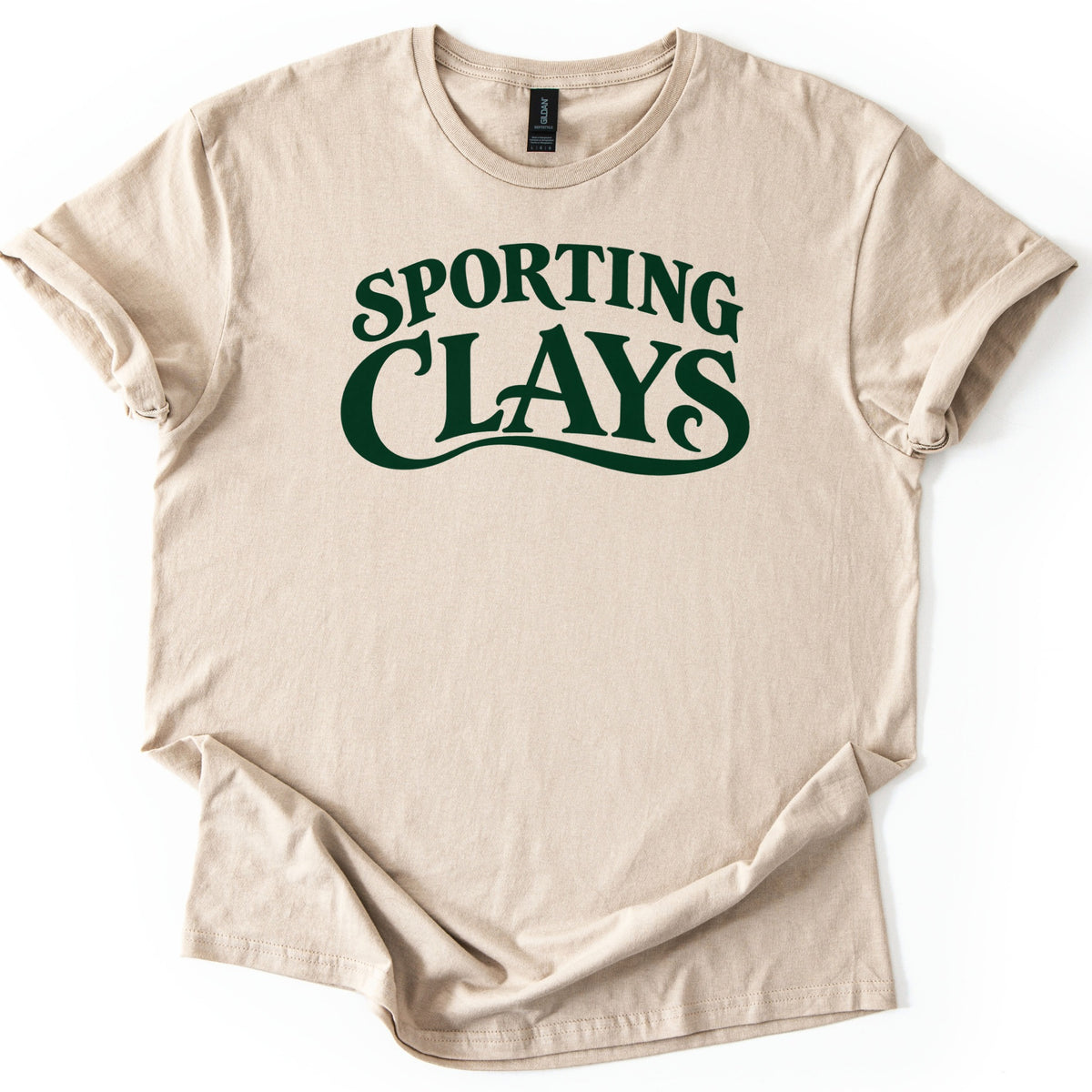 Sporting Clays Comfort Colors T-shirt in Sand, laid flat. Dark green vintage-style lettering reads Sporting Clays, with a curling swash flowing beneath the word Clays. The shirt has rolled sleeves and lies on a plain white background.