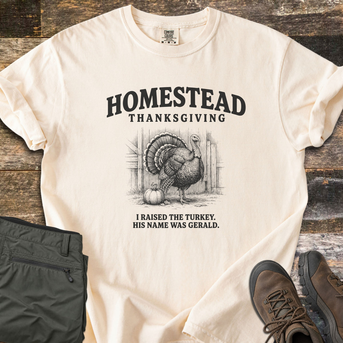 Homestead Thanksgiving Comfort Colors T-shirt in Ivory, laid flat. Black print of a fanned-tail turkey by a barn door and pumpkin, under the words Homestead Thanksgiving, above I Raised the Turkey. His Name Was Gerald. On weathered wood with olive pants and brown hiking boots.