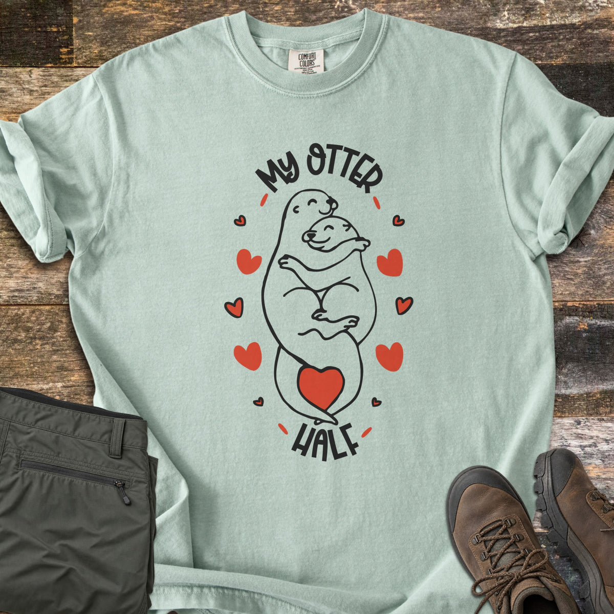 My Otter Half Comfort Colors T-shirt in Bay, laid flat. Two smiling cartoon otters hug in black line art, ringed by red hearts with a red heart at their tails, between the words My Otter Half. Folded olive pants and brown hiking boots sit beside it on weathered wood.