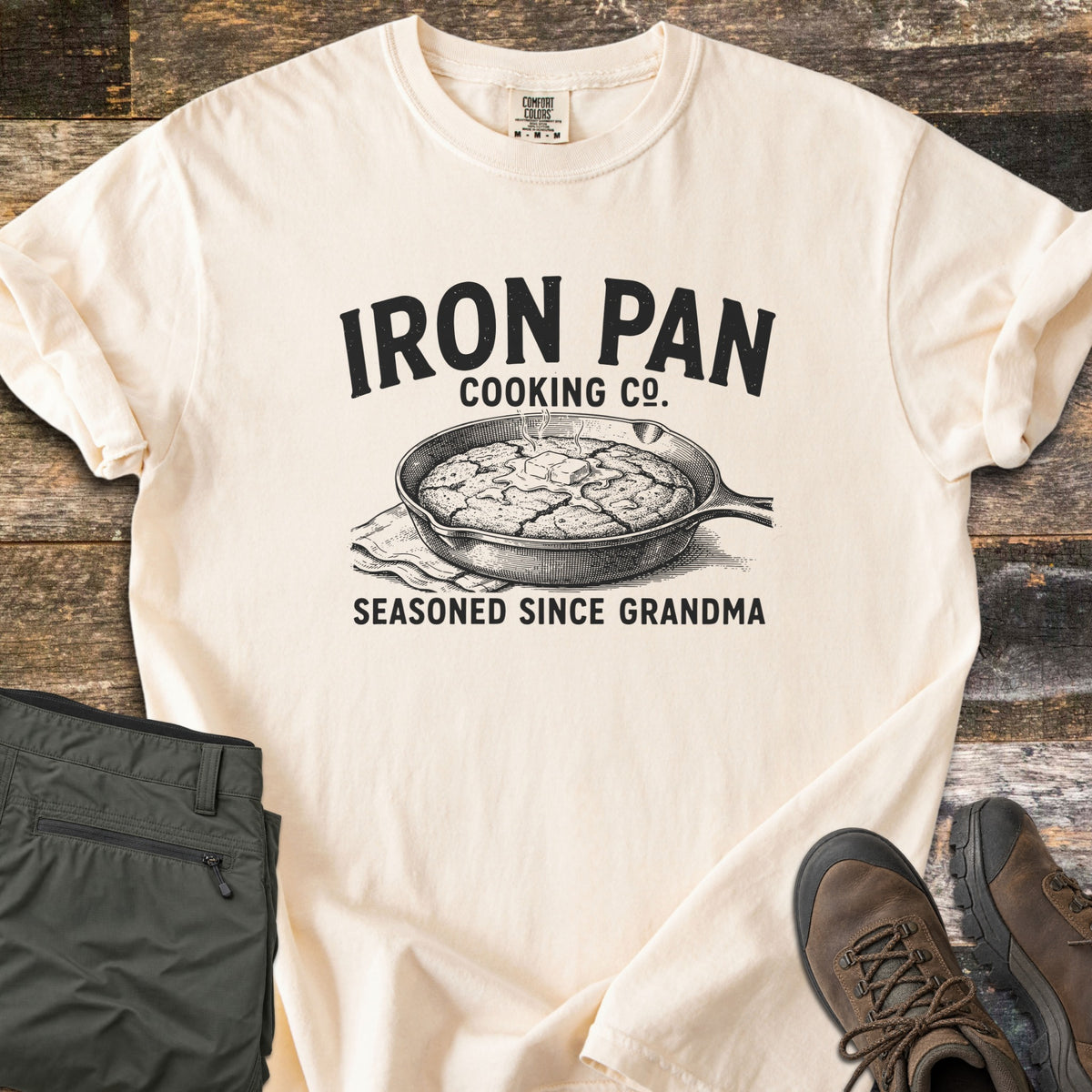 Iron Pan Cooking Co Comfort Colors T-shirt in Ivory, laid flat. Black print of a cast iron skillet of cornbread with melting butter on a napkin, under the words Iron Pan Cooking Co. and above Seasoned Since Grandma. Folded olive pants and brown hiking boots lie on rustic wood.