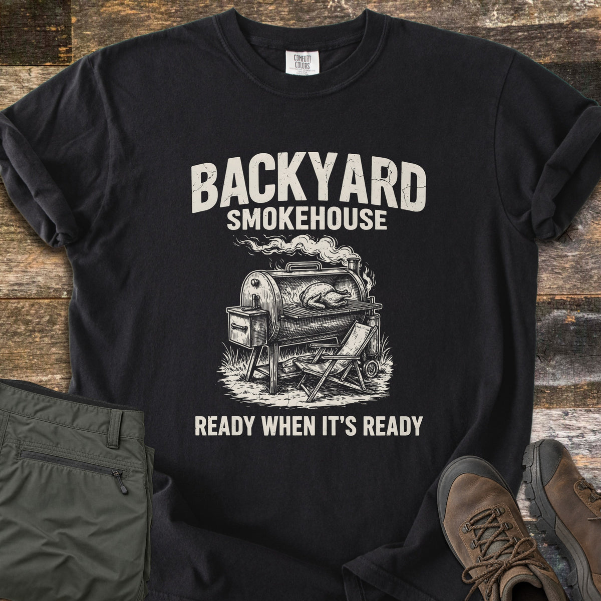 Backyard Smokehouse Comfort Colors T-shirt in Black, laid flat. Cream print of a smoker grill with a chicken inside and a folding chair beside it, under the words Backyard Smokehouse and above Ready When It's Ready. Olive pants and brown boots lie on weathered wood.