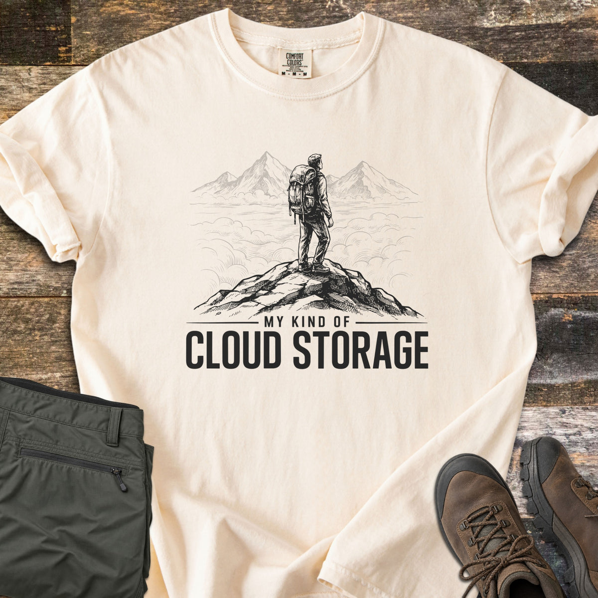 My Kind Of Cloud Storage Comfort Colors T-shirt in Ivory, laid flat. A black sketch of a backpacker on a rocky summit facing peaks above the clouds, over the words My Kind Of Cloud Storage. Folded olive hiking pants and brown hiking boots sit beside it on weathered wood.