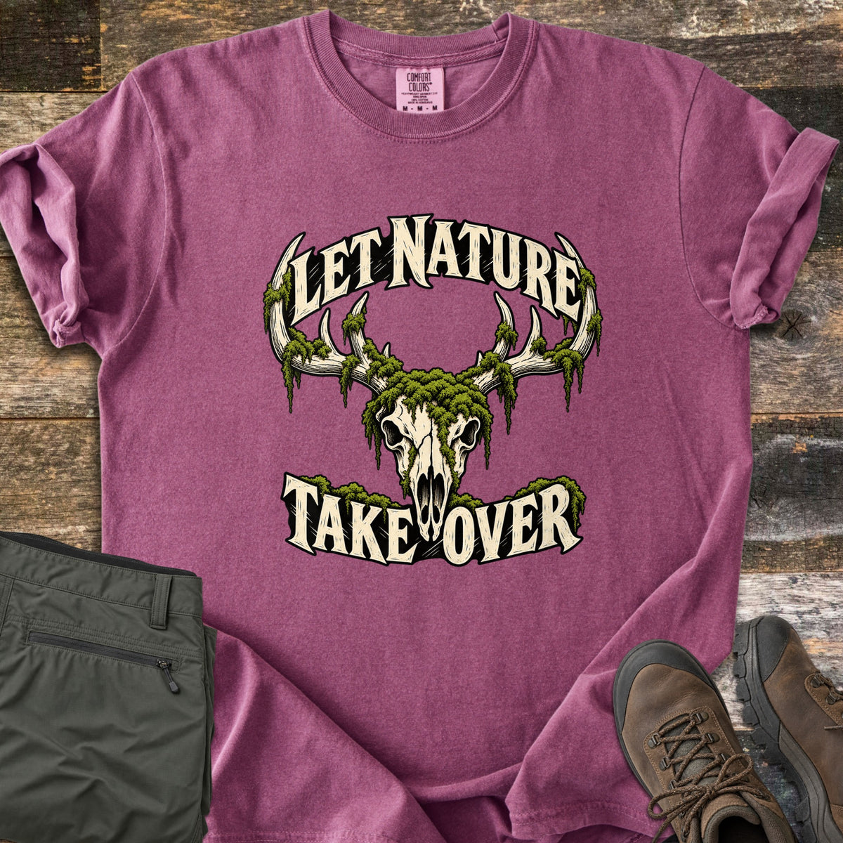 Let Nature Take Over Comfort Colors T-shirt in Berry, laid flat. A moss-covered deer skull with antlers sits between the words Let Nature Take Over in cream lettering. Folded olive hiking pants and brown hiking boots lie beside it on weathered wood planks.