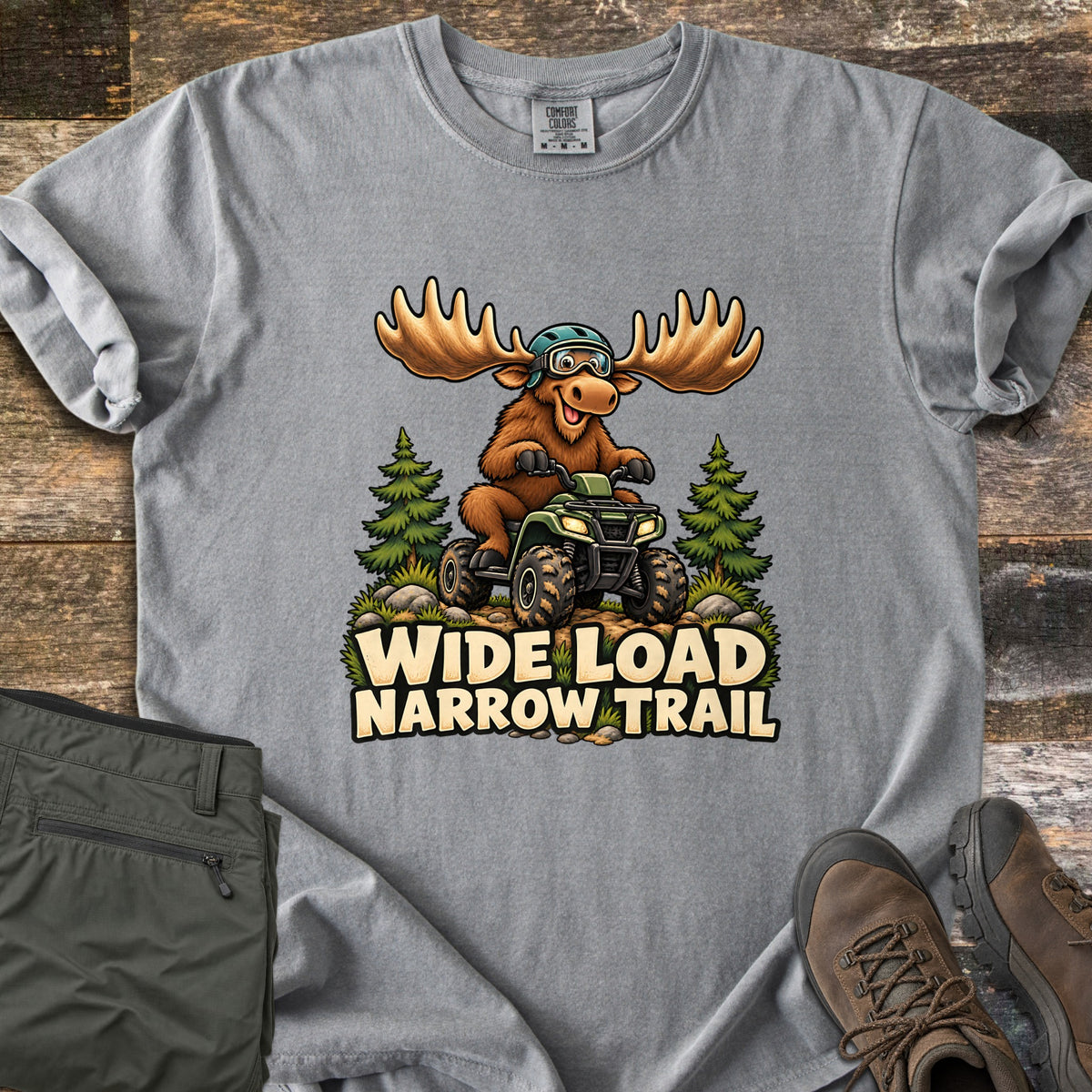 Wide Load Narrow Trail Comfort Colors T-shirt in Grey, laid flat. A cartoon moose with wide antlers, a helmet and goggles rides a muddy green ATV between pine trees above the words Wide Load Narrow Trail. Folded olive pants and brown hiking boots lie on weathered wood.