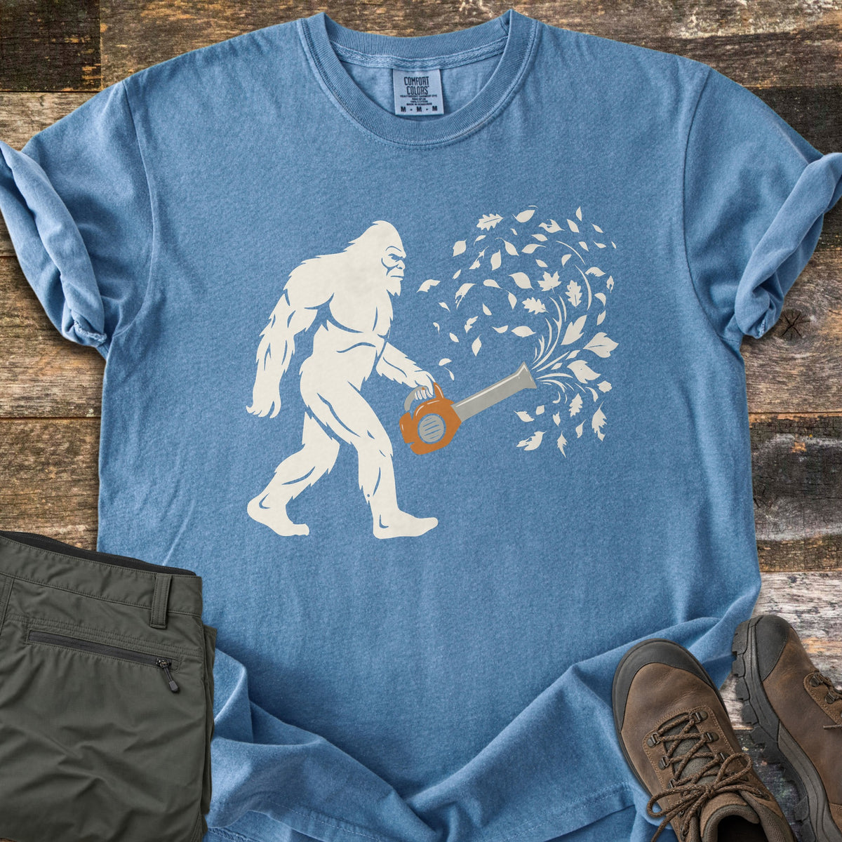 Bigfoot Leaf Blower Comfort Colors T-shirt in Blue Jean, laid flat. A white Bigfoot silhouette walks while holding an orange leaf blower that sends a swirl of white leaves into the air. Folded olive pants and brown hiking boots sit beside it on weathered wood boards.