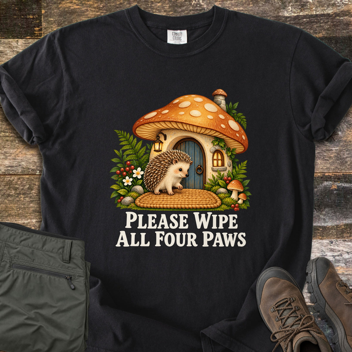 Please Wipe All Four Paws Comfort Colors T-shirt in Black, laid flat. A hedgehog stands on a woven doormat outside a spotted orange mushroom house with a blue door, lantern and ferns, above the words Please Wipe All Four Paws in white. Gray pants and brown hiking boots lie on weathered wood.