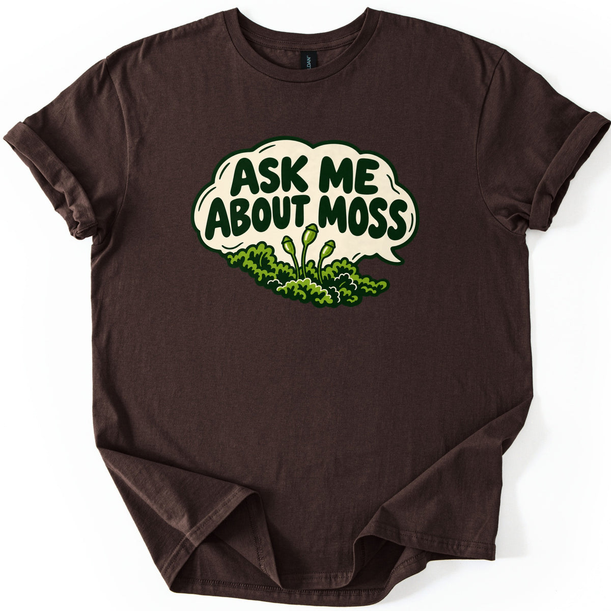 Ask Me About Moss Comfort Colors T-shirt in Dark Chocolate, laid flat. The words ASK ME ABOUT MOSS in bold dark green letters fill a cream speech bubble, with a cartoon patch of green moss and three spore capsules on stalks below. The shirt has rolled sleeves on a white background.