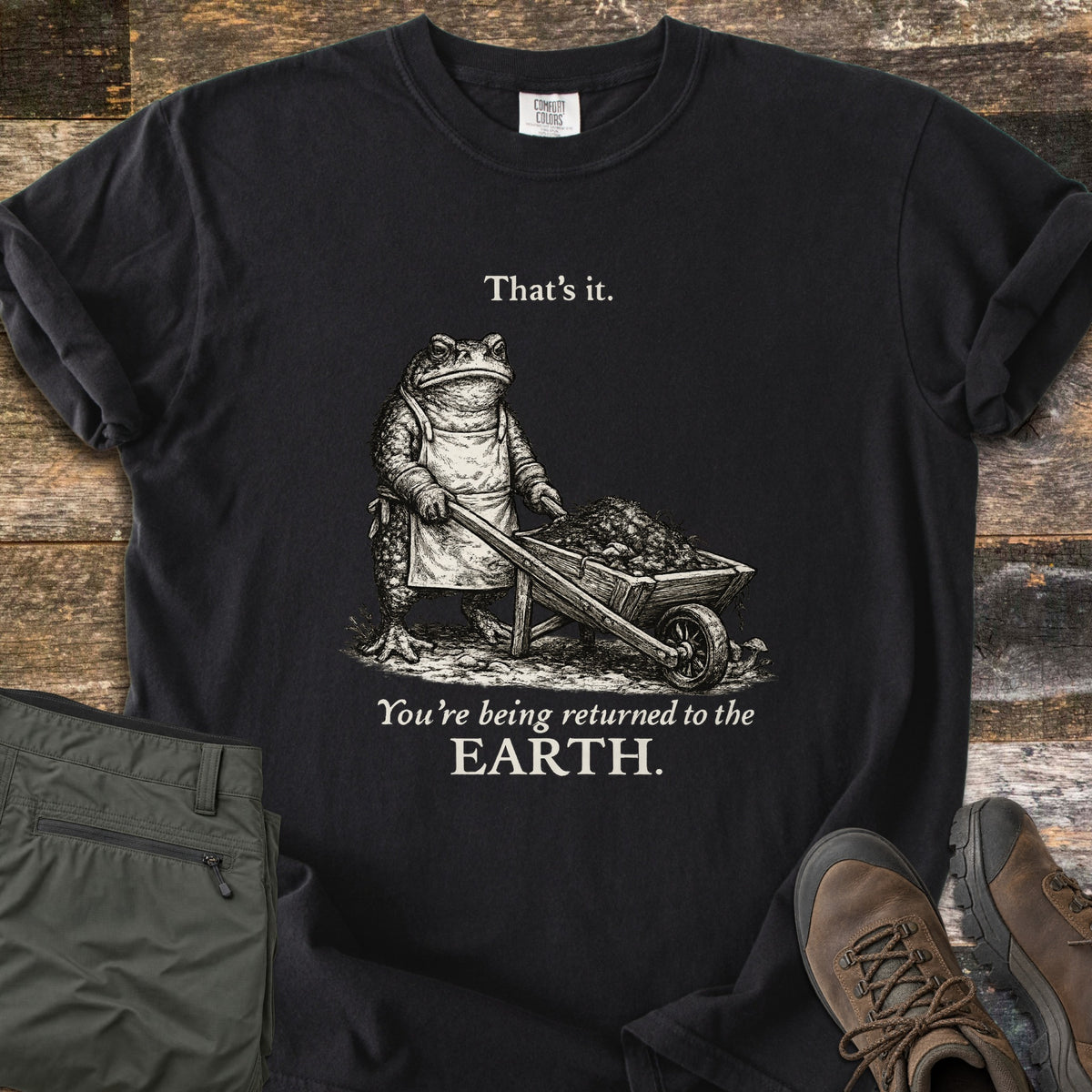 Toad Compost Comfort Colors T-shirt in Black, laid flat. A cream-ink toad in an apron pushes a wheelbarrow of compost between the words That's it. and You're being returned to the EARTH. Folded olive pants and brown hiking boots lie beside it on weathered wood.