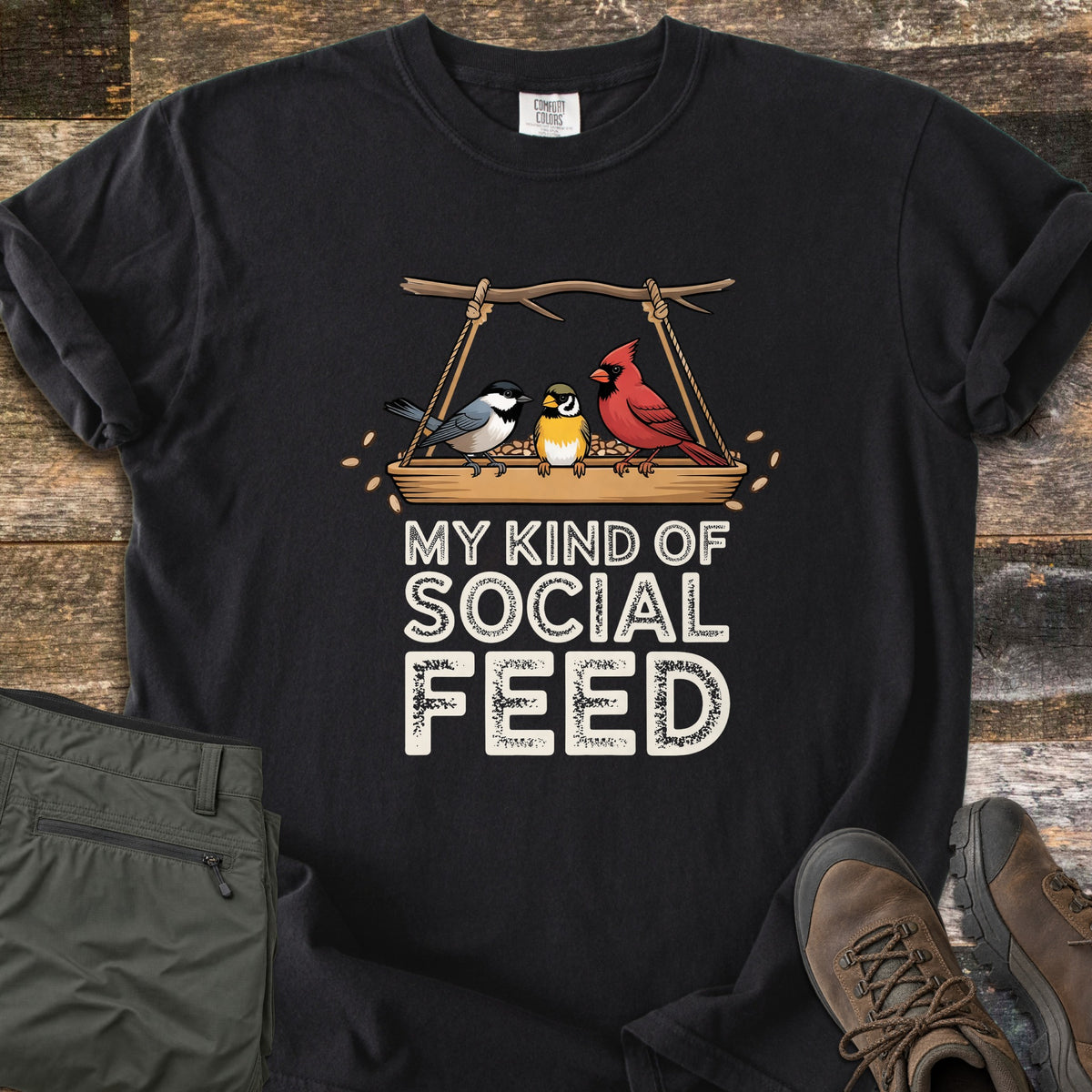 My Kind Of Social Feed Comfort Colors T-shirt in Black, laid flat. A chickadee, a small yellow bird and a red cardinal sit on a rope-hung tray feeder full of seeds above the words My Kind Of Social Feed in white. Olive hiking pants and brown hiking boots lie on weathered wood.