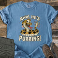 Aww He's Purring Comfort Colors T-shirt in Blue Jean, laid flat. A smiling cartoon rattlesnake with diamond markings shakes its rattle, framed by the words Aww, He's Purring! The shirt has rolled sleeves and lies on weathered wood with olive pants and brown hiking boots.
