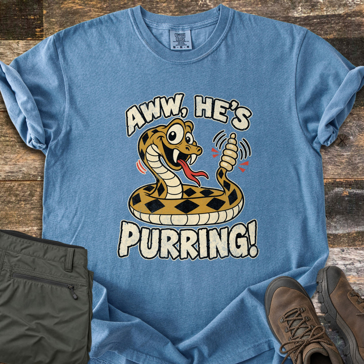 Aww He's Purring Comfort Colors T-shirt in Blue Jean, laid flat. A smiling cartoon rattlesnake with diamond markings shakes its rattle, framed by the words Aww, He's Purring! The shirt has rolled sleeves and lies on weathered wood with olive pants and brown hiking boots.