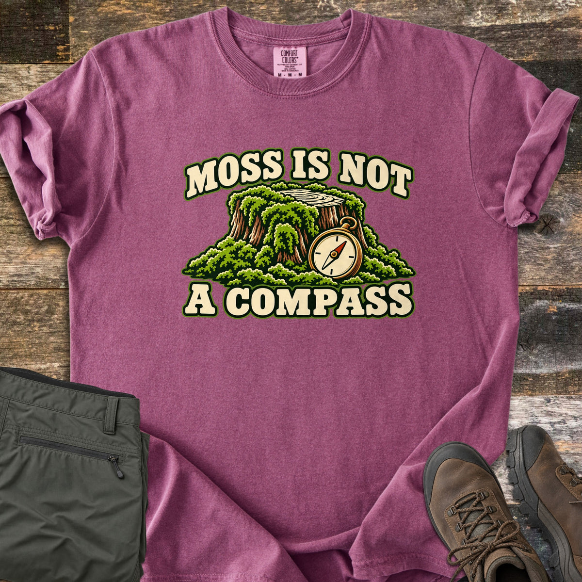 Moss Is Not a Compass Comfort Colors T-shirt in Berry, laid flat. Printed is a mossy tree stump with a brass compass resting against it, under MOSS IS NOT and above A COMPASS. Folded olive hiking pants and brown boots rest on old wood boards.