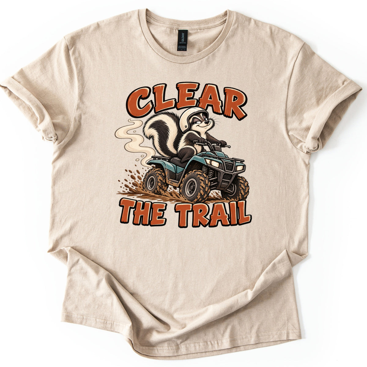 Clear the Trail Comfort Colors T-shirt in Sand, laid flat. A grinning cartoon skunk in a white helmet rides a muddy teal ATV trailing smoke, with Clear above and The Trail below in rust-orange letters. The shirt lies on a white background with cuffed sleeves.