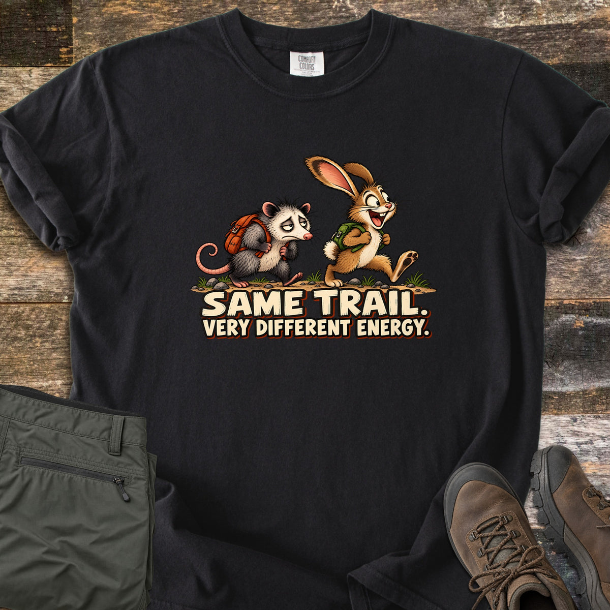 Same Trail. Very Different Energy. Comfort Colors T-shirt in Black, laid flat. A tired cartoon opossum with an orange backpack trudges behind a grinning, bounding rabbit with a green backpack on a dirt trail, above the words Same Trail. Very Different Energy. Shown on wood with green pants and hiking boots.