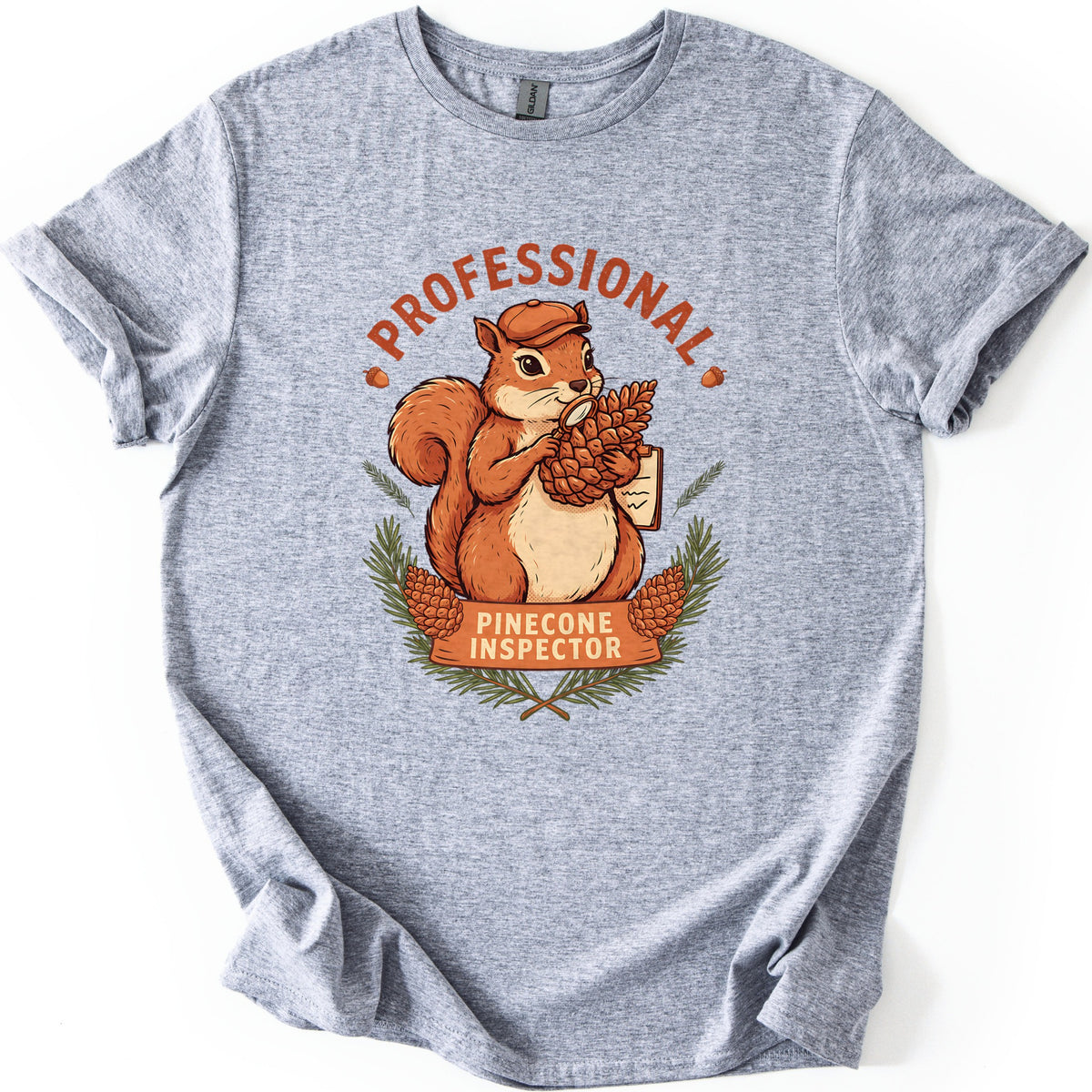Professional Pinecone Inspector Comfort Colors T-shirt in Sport Grey, laid flat. A squirrel in a cap inspects a pinecone with a magnifying glass under the arched word Professional in rust red, above a banner reading Pinecone Inspector framed by pine branches. Sleeves are cuffed on a white surface.