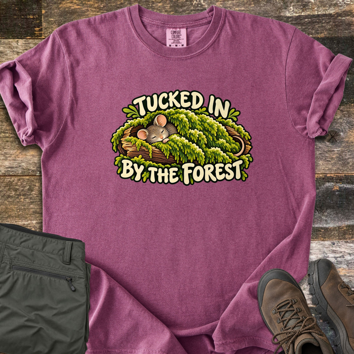 Tucked in by the Forest Comfort Colors T-shirt in Berry, laid flat. A sleeping cartoon mouse lies under a blanket of green moss in a hollow log between the words Tucked In By The Forest. Folded olive hiking pants and brown hiking boots sit on weathered wood planks.